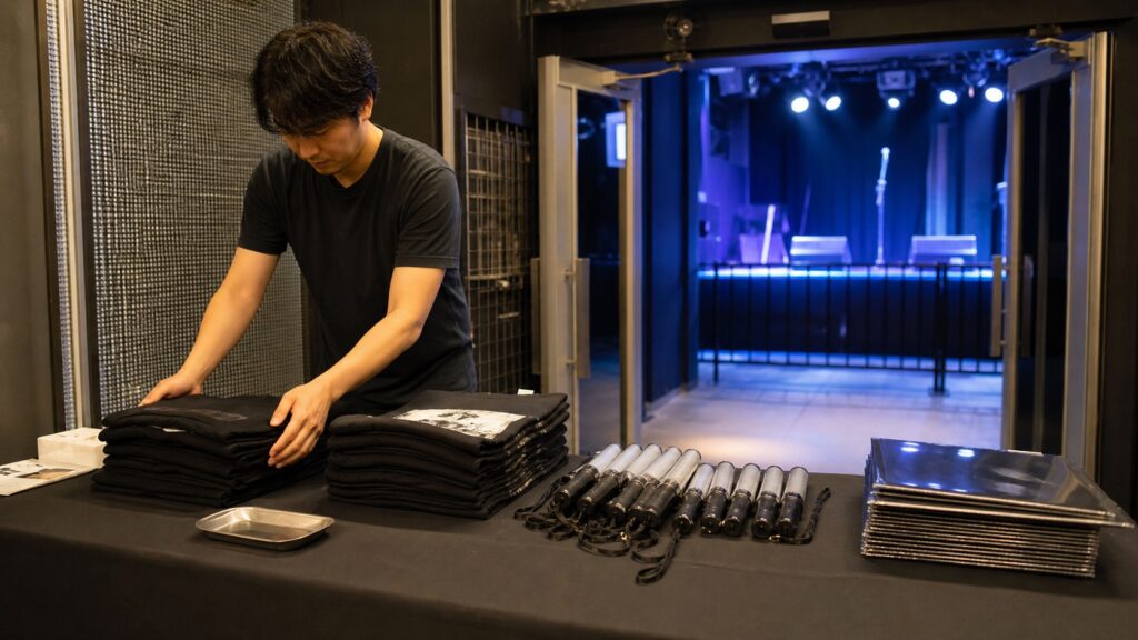 小さなライブハウスの物販卓でグッズを準備する架空の成人スタッフ