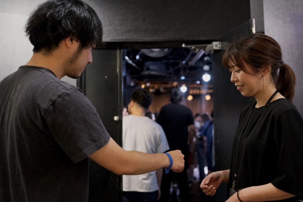 架空のライブ会場でスタッフに無地のリストバンドを見せる来場者
