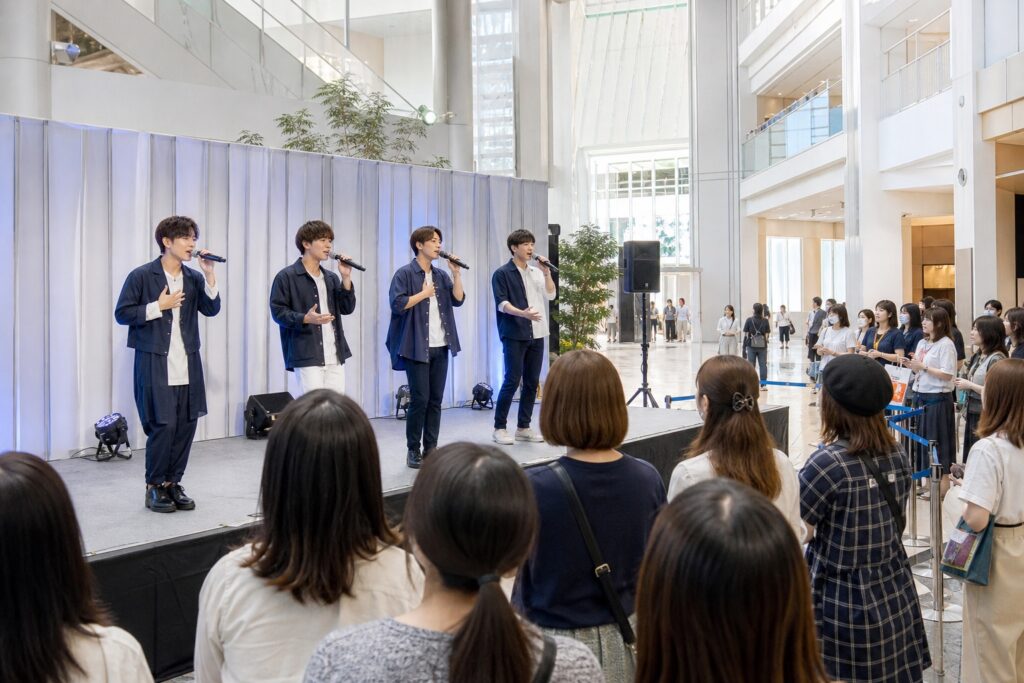 架空の商業施設の小さなステージで歌う男性アイドルと観客